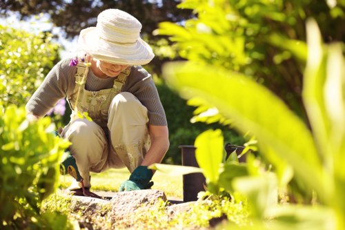 A woman wearing a wide-brimmed sun hat, beige gardening apron, and gloves is kneeling in a well-maintained garden in Tooting, tending to a flower bed with her hands. The garden features lush green grass, neatly edged flower beds, and a variety of shrubs and small plants. In the background, there are trees and additional garden borders, with sunlight filtering through the foliage, creating a warm and bright outdoor environment. The scene captures a peaceful moment of gardening maintenance, with a focus on outdoor care and landscaping. The setting is indicative of a typical suburban garden in Tooting, suitable for lawn care, hedge trimming, and planting services offered by Lawn Mowing Tooting, reflecting a natural and inviting outdoor space.