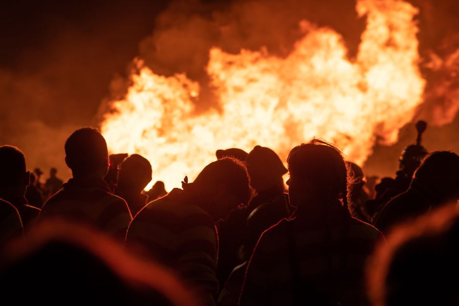 A large bonfire burning intensely during the night, with bright orange flames engulfing a pile of wooden logs and debris. The flames cast a warm glow against the dark sky, revealing textures of charre